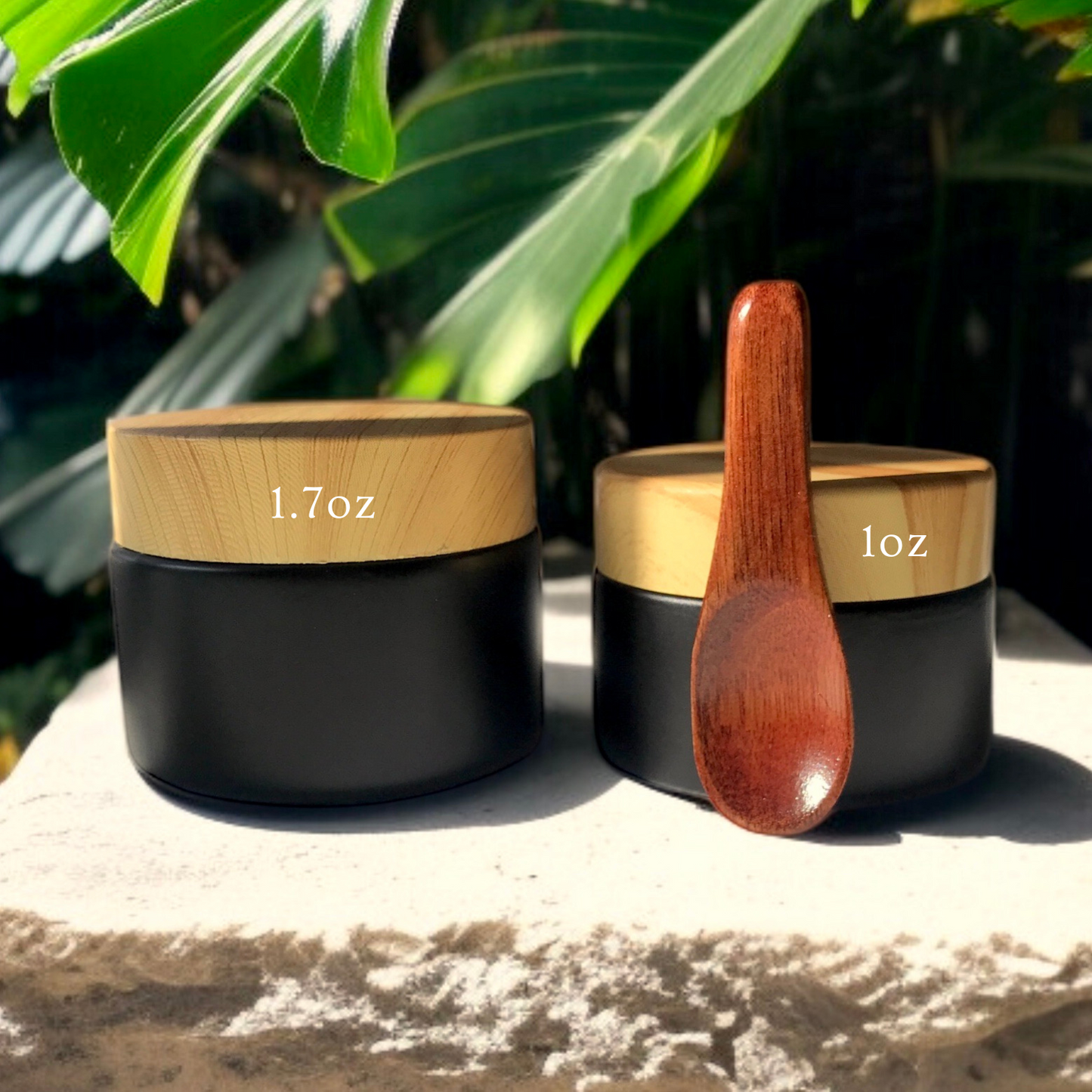 Two black containers with wooden lids and a wooden spoon on a stone surface with green foliage in the background.