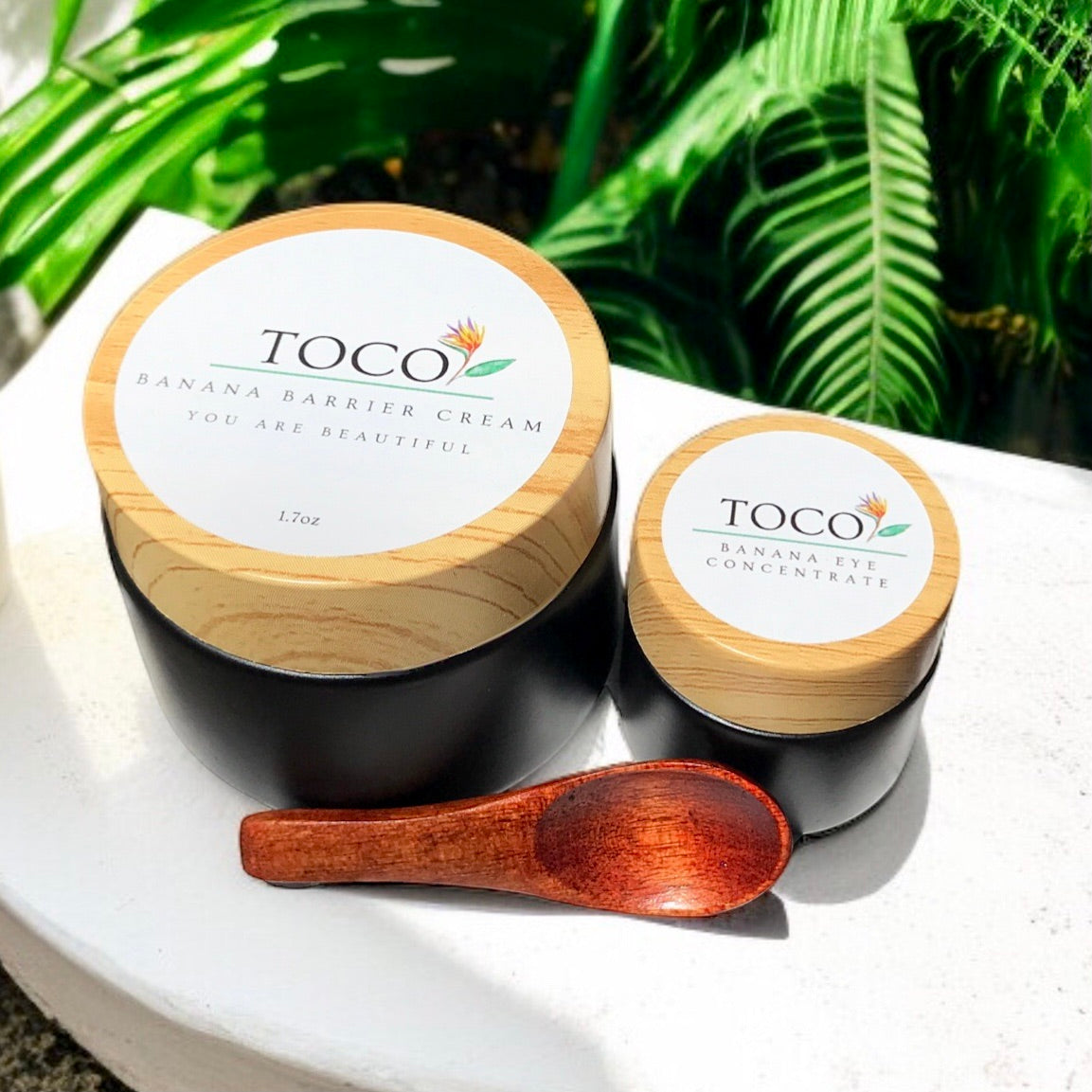 Two Toco skincare products with wooden lids and a spoon on a white surface with green leaves in the background.