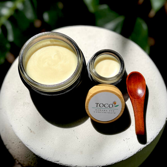 Banana Barrier Cream and Banana Eye Concentrate with a small wooden spoon on stone surface with tropical plants
